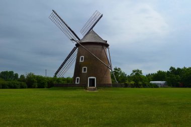 Tuğla temeli ve ahşap bıçakları olan geleneksel bir yel değirmeni ağaçlarla çevrili çimenli bir tarlada duruyor ve bulutlu bir gökyüzünün altında küçük bir yapı..