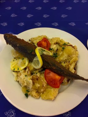 Limon ve maydanozla süslenmiş, domates dilimli kremalı patates salatasıyla servis edilmiş, mavi çiçekli bir masa örtüsünün üstünde tam kaplama balık..