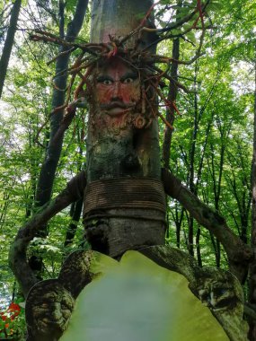 Yeşil yapraklar ve ek sanat eserleri ile çevrili, bir ormanda boyanmış bir yüz ve ip gibi bir insan heykeli duruyor..