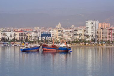 İki rıhtımlı geminin, palmiye ağaçlarıyla dolu bir gezinti güvertesi, orta ve yüksek binalar ve arka planda puslu dağların yer aldığı bir kıyı şehri..