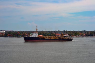 Karanlık ve beyaz gövdeli bir kargo gemisi durgun sularda duruyor, binalar, palmiye ağaçları ve parçalı bulutlu gökyüzü ile yeşil bir kıyıya kurulmuş..