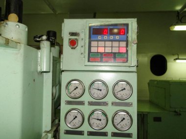 Dijital ekranlı endüstriyel kontrol paneli, altı basınç göstergesi ve lomboz pencereli açık yeşil motor odasında kırmızı acil durum düğmesi..