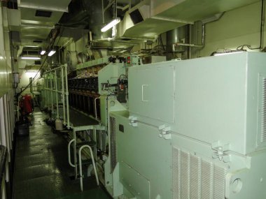 Büyük bir deniz mazotu motoru, yeşil kayma önleyici döşeme, kontrol panelleri, borular ve kırmızı yangın söndürücünün yer aldığı iyi aydınlatılmış gemi makine dairesi..
