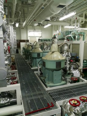 Modern bir tesiste yeşil santrifüj ayırıcıları, rendelenmiş metal geçitler, üstten borular, floresan aydınlatma ve kontrol panelleri bulunan makine dairesi..