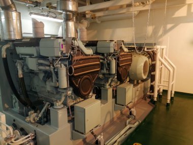 İki büyük gri endüstriyel motorun, egzoz borularının, havalandırma borularının, yeşil zeminin, organize boruların ve kabloların bulunduğu mekanik oda..