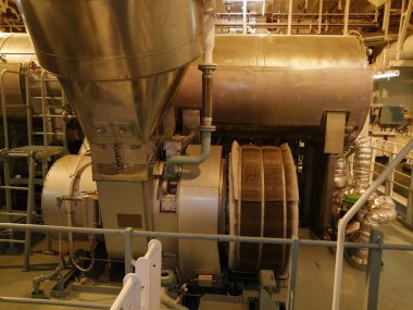 Silindirik varili, boruları, yalıtılmış bölümleri ve rendelenmiş bir yürüyüş yolu olan büyük metalik bir makinenin bulunduğu temiz ve işlevsel bir makine dairesi..