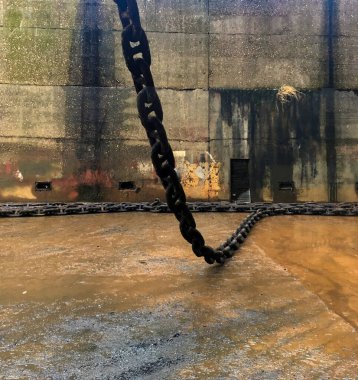 Large chain partially on wet ground in a dry dock, with a weathered concrete wall showing rust, stains, and small rectangular openings. clipart