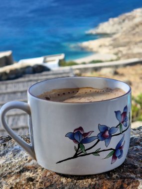 Üzerinde çiçek deseni olan bir fincan kahve okyanusa bakan bir kayanın üzerinde duruyor.