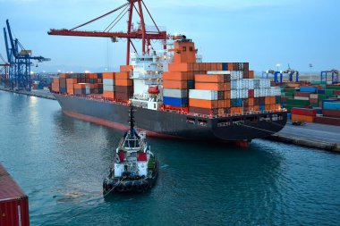 Römorkörle birlikte limana yanaşmış renkli konteynırları olan büyük bir kargo gemisi. Turnalar ve konteyner yığınları deniz lojistiğini vurguluyor.