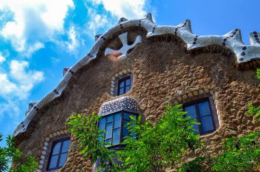 İspanya, Barselona, Park Guell 'de seramik kiremitleri, mozaik çerçeveli pencereleri ve mavi gökyüzü altında yemyeşil yaprakları olan bir bina..