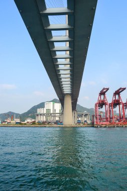 Köprünün su altındaki görüntüsü, merkezi destek kolonu, kırmızı nakliye vinçleri, şehir binaları, yeşil tepeler ve açık mavi gökyüzü..