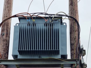 İki ahşap elektrik direğine monte edilmiş soğutucu yüzgeçleri olan gri elektrik trafosu, bulutlu bir gökyüzünün altında renkli kablolarla birbirine bağlı..