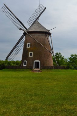 Tuğla tabanlı, ahşap bıçaklı, iyi korunmuş bir yel değirmeni ve parçalı bulutlu bir gökyüzünün altında çimenli bir arazide geçen çitli bir çevre..