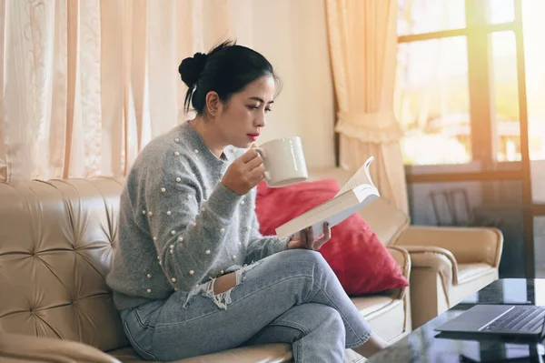 woman relaxing at home , woman reading book and drink coffee holding a cup of coffee sitting on sofa - relaxing in her living room reading book on holiday is favorite hobby in home and comfort