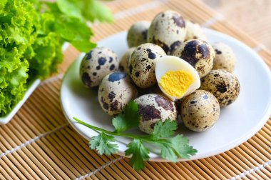 Haşlanmış yumurta, beyaz tabakta bıldırcın yumurtası, taze bıldırcın yumurtalı kahvaltı yumurtası ve ahşap masa arkasında sebze kişniş.