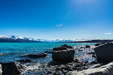 Göz kamaştırıcı bir göl manzarası berrak su, kayalar ve parlak gökyüzünün altında görkemli dağlar sergiliyor..