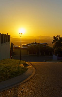 Gün sona ererken, güneş güzel bir şekilde huzurlu bir yerleşim alanının üzerinde batıyor. Önü aydınlatan bir sokak lambası var.