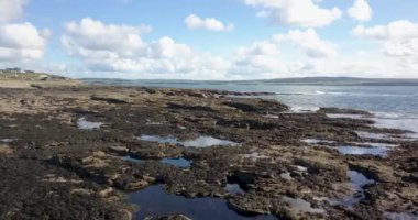 Bulutlu bir gökyüzünün altındaki sakin suların yanında engebeli kayalık bir kıyı şeridinin çarpıcı ve pitoresk bir görüntüsü.