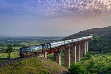 Pune, Maharashtra, India-October 8th, 2017: Along with Sunrise, Indian railways long passenger train, exiting a tunnel to cross a tall railway bridge. clipart
