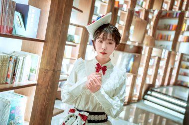 Bu resim, bir kütüphanenin içinde bulanık bir kitaplık arka planı olan beyaz elbiseli genç bir kadını gösteriyor..