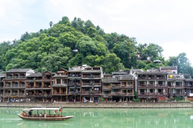 English description: Traditional Miao architecture by the river in Fenghuang Ancient Town, surrounded by greenery and hills. clipart