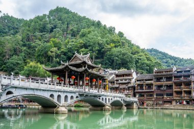 English description: Traditional Miao architecture by the river in Fenghuang Ancient Town, surrounded by greenery and hills. clipart