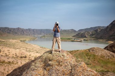 Professional photographer takes photos with camera in hands on rocky peak. Look at camera, cipy space.