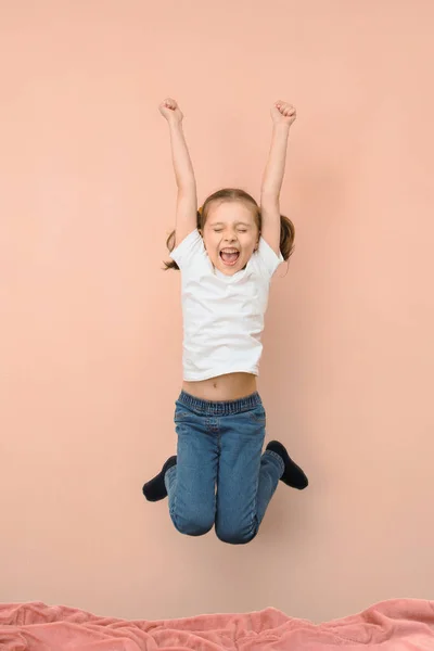 A little girl in a jump on a bed screams with happiness on a pink background.