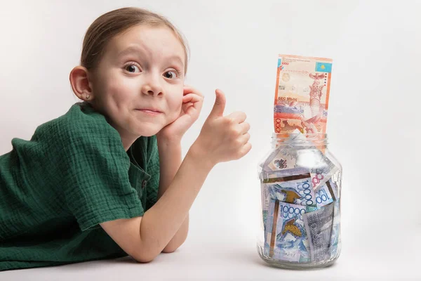 Happy thumb up girl with a large transparent jar with Qazaqstan tenge money. Bank deposit for a child.