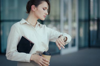 Elinde not defteri, elinde kahve bardağı olan bir iş kadını saatine bakıyor. Modern hızda zaman yönetimi kavramı.
