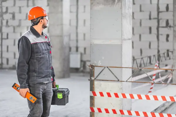A foreman inspects a new apartment before starting work. Builder in uniform carrying toolbox and level tool at construction site, copy space.