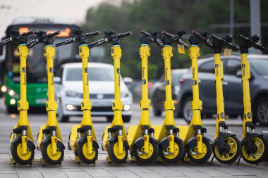 Yandex 'ten kiralanmaya hazır birçok elektrikli scooter, otomobil ve otobüslerin yoğun trafiğiyle bir otoyolun arka planında ayakta duruyor. Almaty, Kazakistan - 11 Haziran 2023