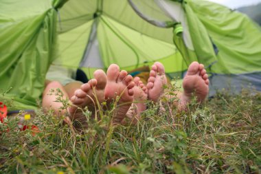 İki çift çıplak ayak, doğanın tadını çıkarırken çimenli bir çayıra kurulmuş bir çadırda rahatça dinlenir.