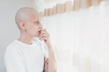 Kemoterapiden sonra saçlarını kaybeden beyaz tişörtlü genç bir kadın pencerenin kenarında duruyor, kameraya bakıyor, boşluğu kopyalıyor..