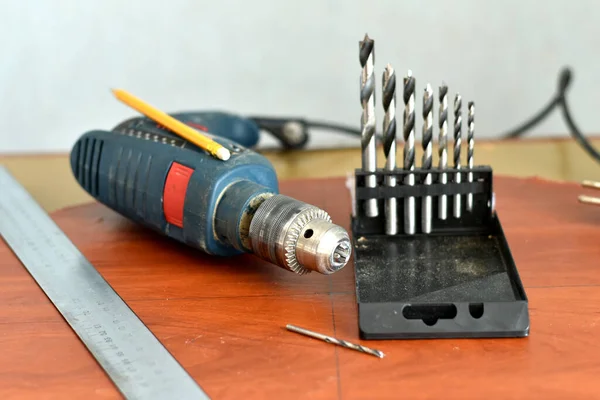 In the picture, a drill, drill bits and a metal ruler are on the table.