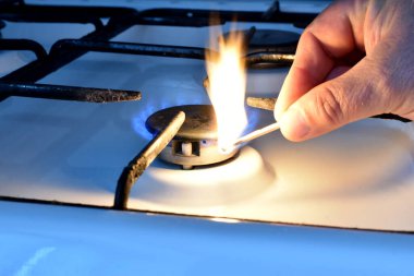 Resim bir adamın elini gösteriyor. Bir adam elinde bir kibrit tutuyor ve onu gaz sobasının ocağına getiriyor..