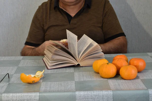 Mężczyzna Siedzi Przy Stole Przed Otwartą Księgą Obiera Mandarynkę — Zdjęcie stockowe