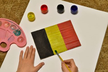 The flag of Belgium is drawn by a boy on a piece of paper with watercolors and a brush.