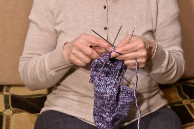 Women's hands hold knitting needles with which a woman knits a woolen sock.