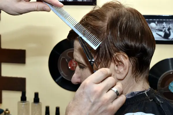 A hairdresser using a comb and scissors shortens the hair on the temples of a womans head, side view.