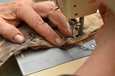 Resim, dikiş makinesi kullanarak iki parça kumaşı bir araya getiren bir terzinin elini gösteriyor..