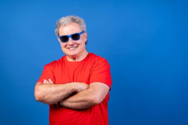 Kırmızı gömlekli bir adam gülümsüyor ve güneş gözlüğü takıyor. Mavi arka planda bir resim için poz veriyor.