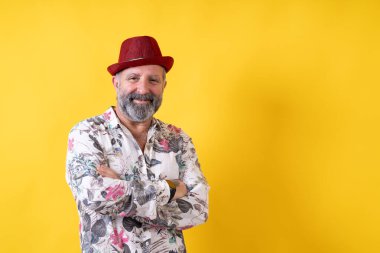 Kırmızı şapkalı ve çiçekli gömlek giyen bir adam. Gülümsüyor ve kollarını kavuşturuyor.