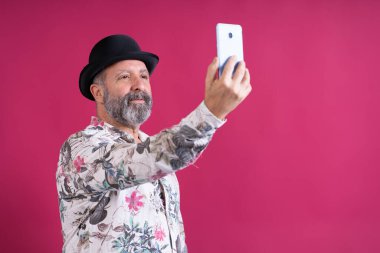 Bir adam cep telefonuyla kendi fotoğrafını çekiyor. Siyah bir şapka ve çiçekli bir gömlek giyiyor.