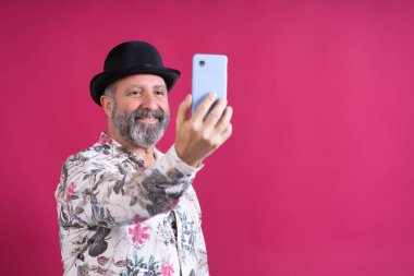 Adamın biri cep telefonuyla selfie çekiyor. Siyah bir şapka ve çiçekli bir gömlek giyiyor.