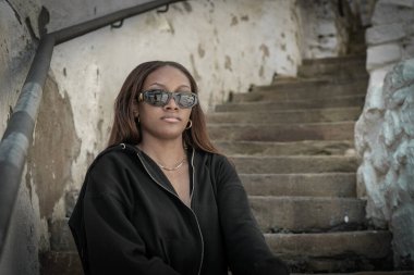 Güneş gözlüğü takan ve siyah kapüşonlu bir kadın merdivende oturuyor. Gizem ve entrika kavramı, kadının güneş gözlüğü ve merdiven ayarları gizlilik ortamı yaratıyor.