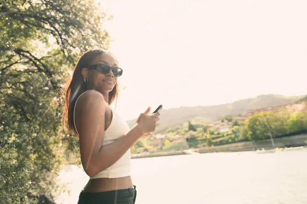 Bir kadın su birikintisinin yanında duruyor, elinde cep telefonu var. Gülümsüyor ve dışarıda eğleniyor.