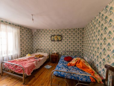 Terk edilmiş bir evin yatak odasında iki adet yapılmamış metal yatak duruyor. Eski duvar kâğıdı sergileniyor ve firar hissi uyandırıyor.