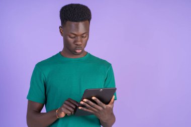 Mor arka planda tablet kullanan yeşil tişört giyen genç adam yoğun bir şekilde bir şeyler okuyor.