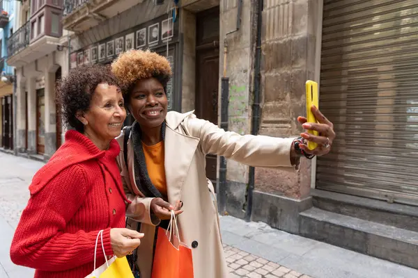 İki neşeli çoklu etnik kadın akıllı bir telefonla selfie çekiyor, canlı bir şehir caddesinde alışveriş çılgınlıklarını kutluyorlar.